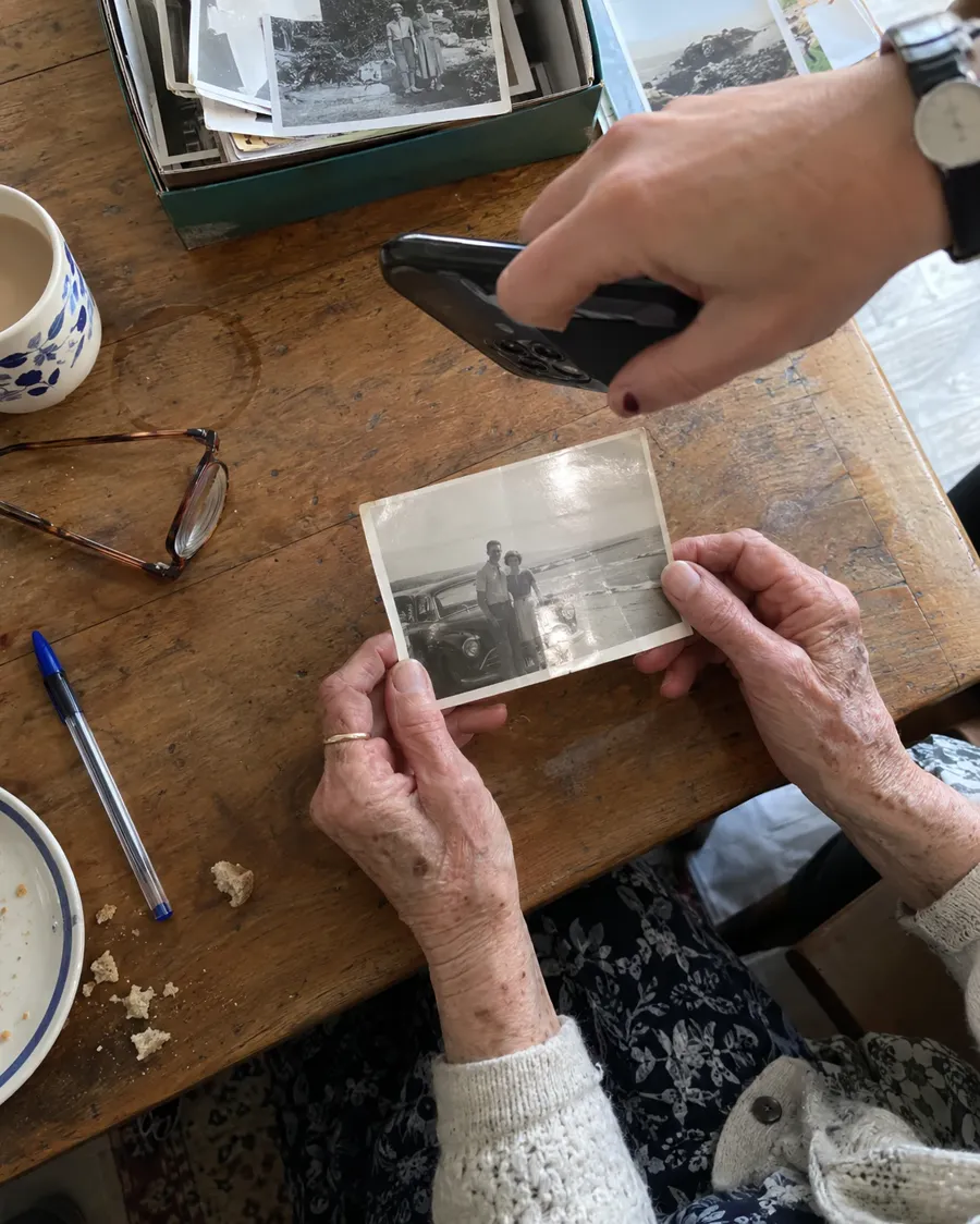 Older hands holding an old black-and-white beach photo while someone photographs it with a phone